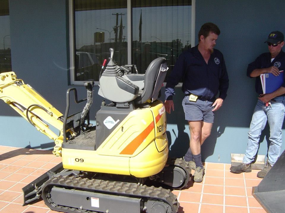 Yellow mini excavator with two men standing nearby. One man is wearing shorts and a blue shirt.