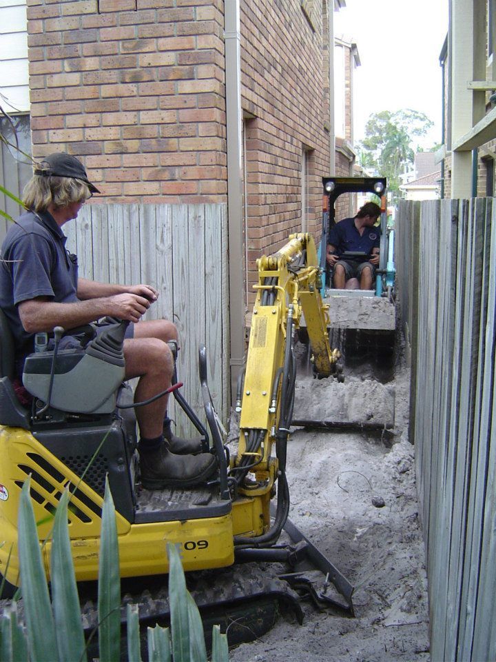 Two people using excavators to dig in a narrow space between a brick building and a wooden fence.