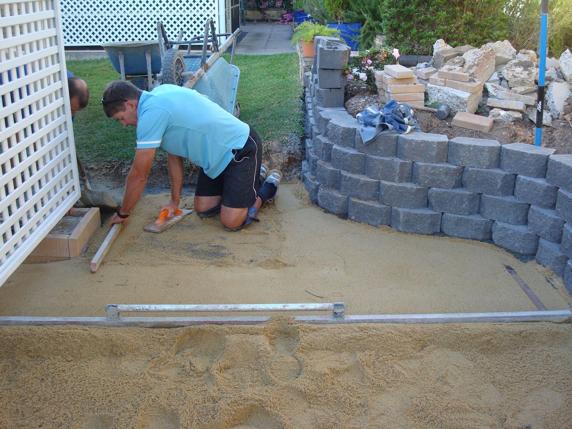 Man kneeling, leveling sand for paving stones next to a retaining wall.