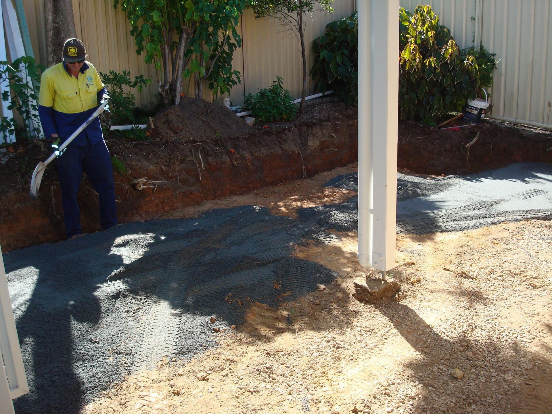 Construction worker spreading gravel with a shovel near a white support column and prepared ground.