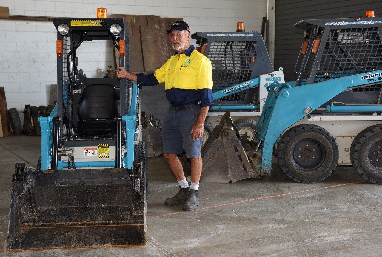 Man in work clothes standing by a small, blue skid-steer loader inside a building.