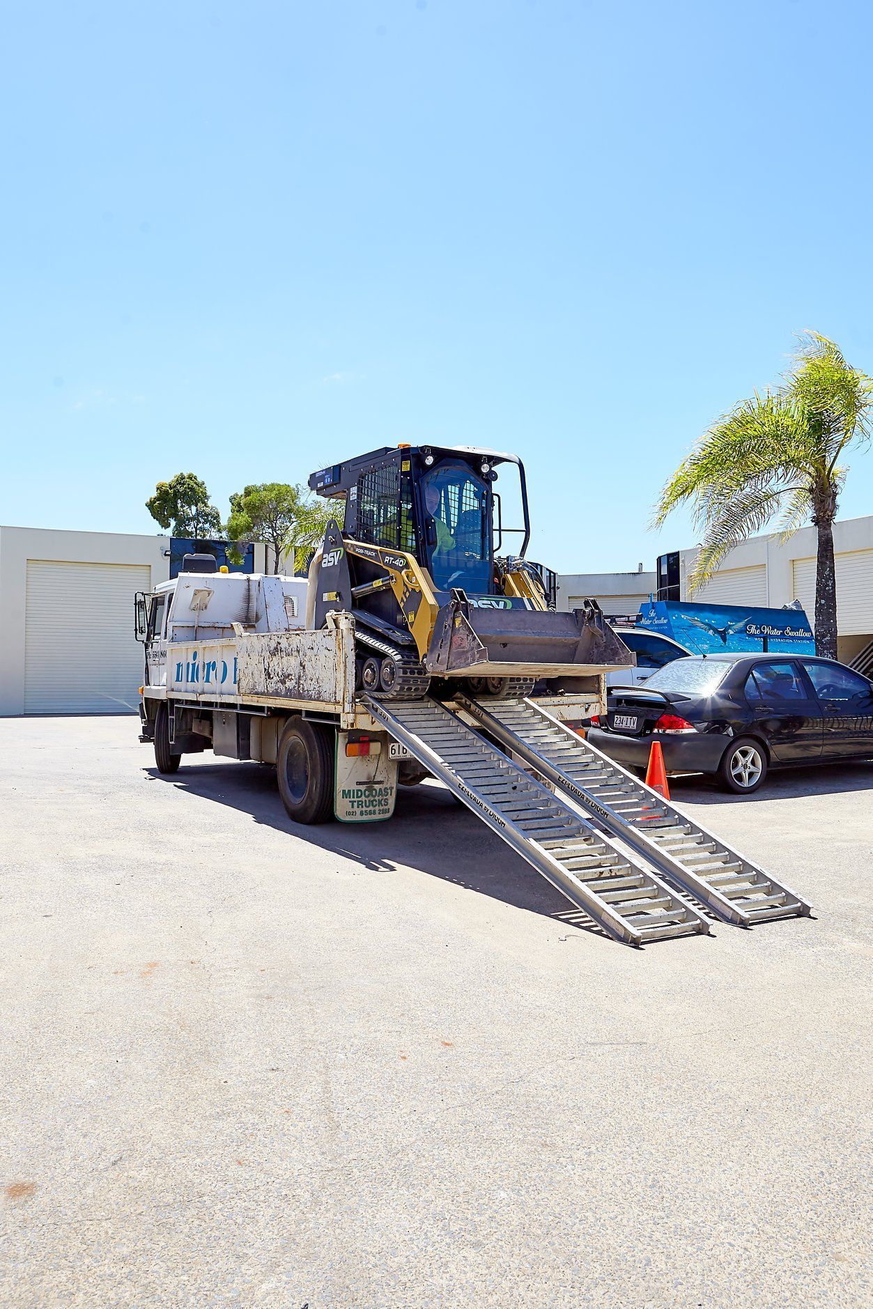 A small construction vehicle on a truck bed with ramps, parked outside on a sunny day.