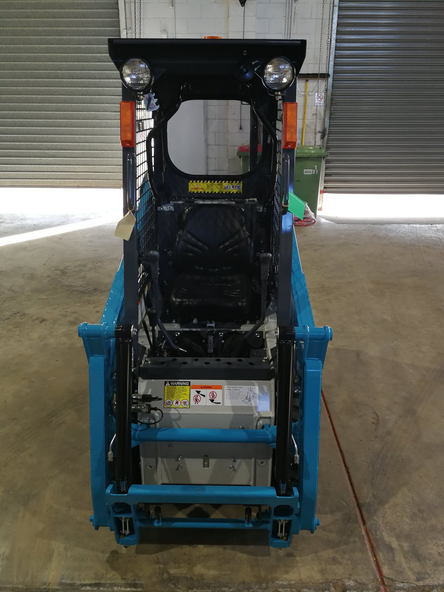 Blue skid steer loader inside a garage, with overhead lights and orange side markers.