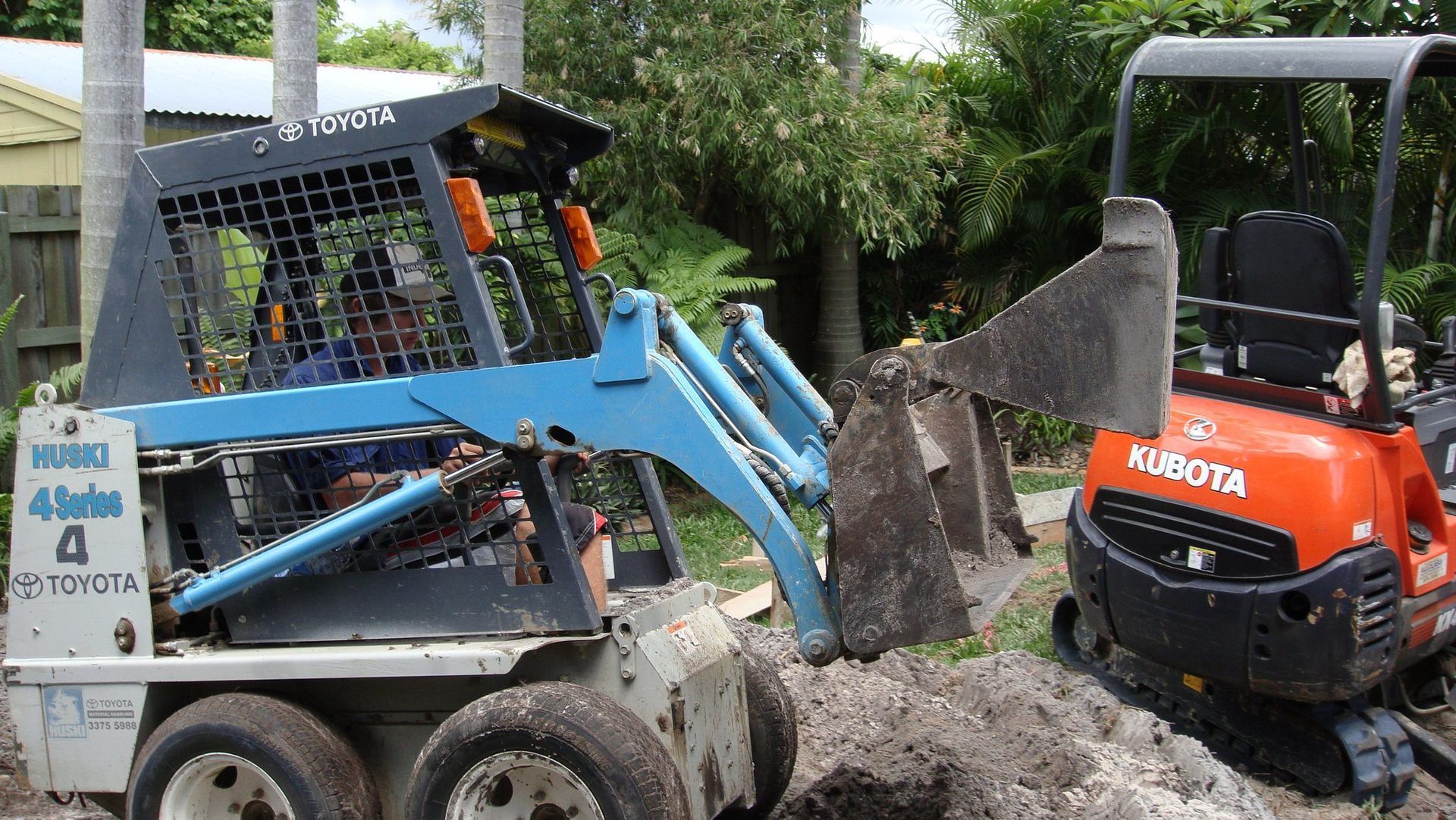Blue skid steer loader and orange Kubota excavator digging in a yard.
