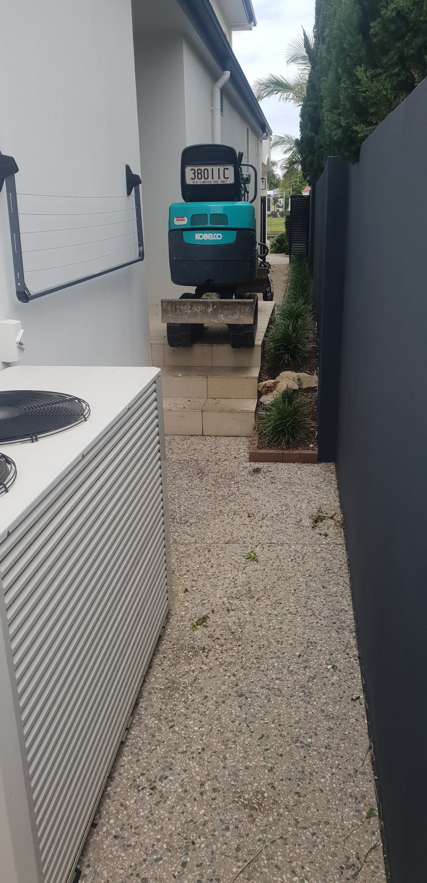 A small excavator sits between a building and a black fence on a gravel pathway.