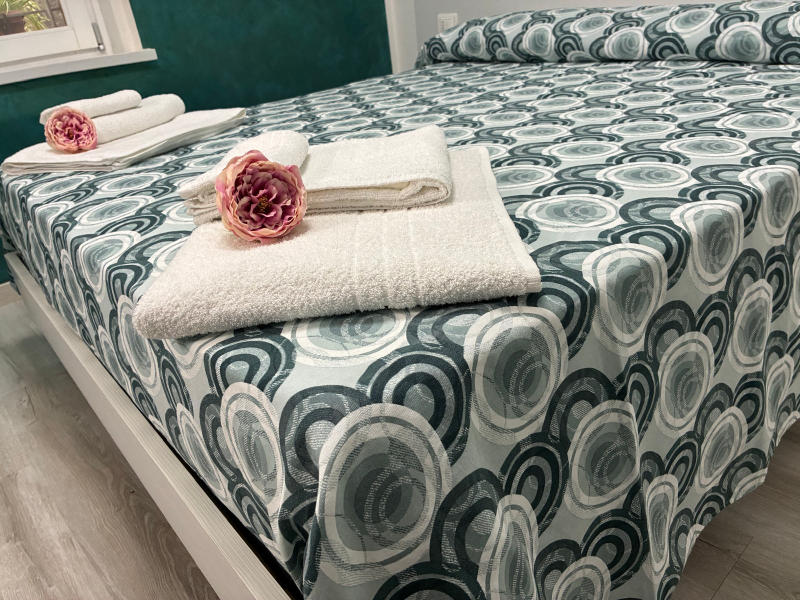 Letto ben rifatto con copriletto a fantasia color verde acqua e asciugamani bianchi arrotolati, sormontati da fiori rosa.