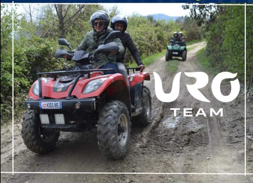 Due persone a bordo di un quad rosso su un sentiero fangoso, con un altro quad alle loro spalle e la scritta