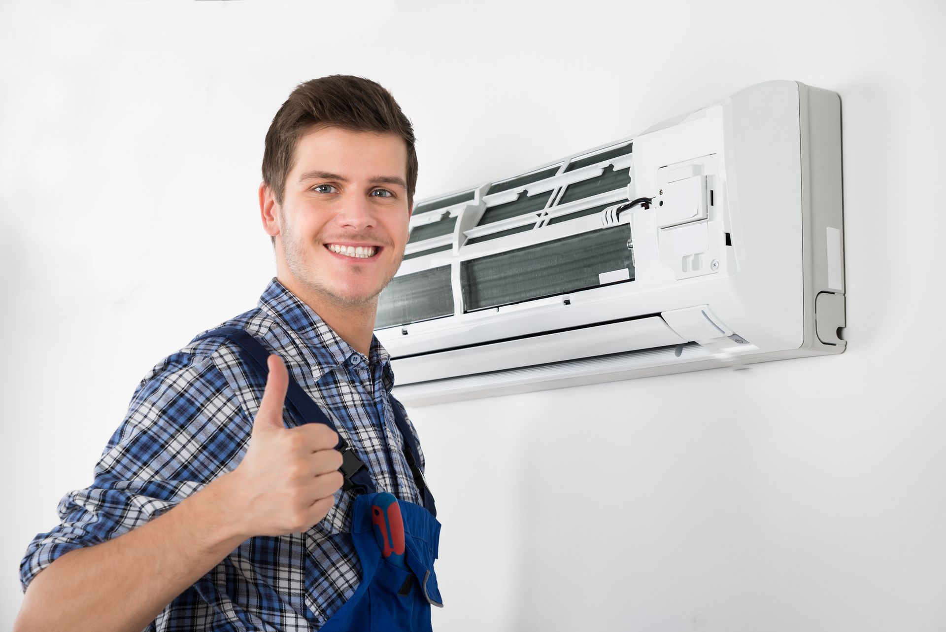 Man in work uniform gives thumbs up near an air conditioner unit on a white wall.