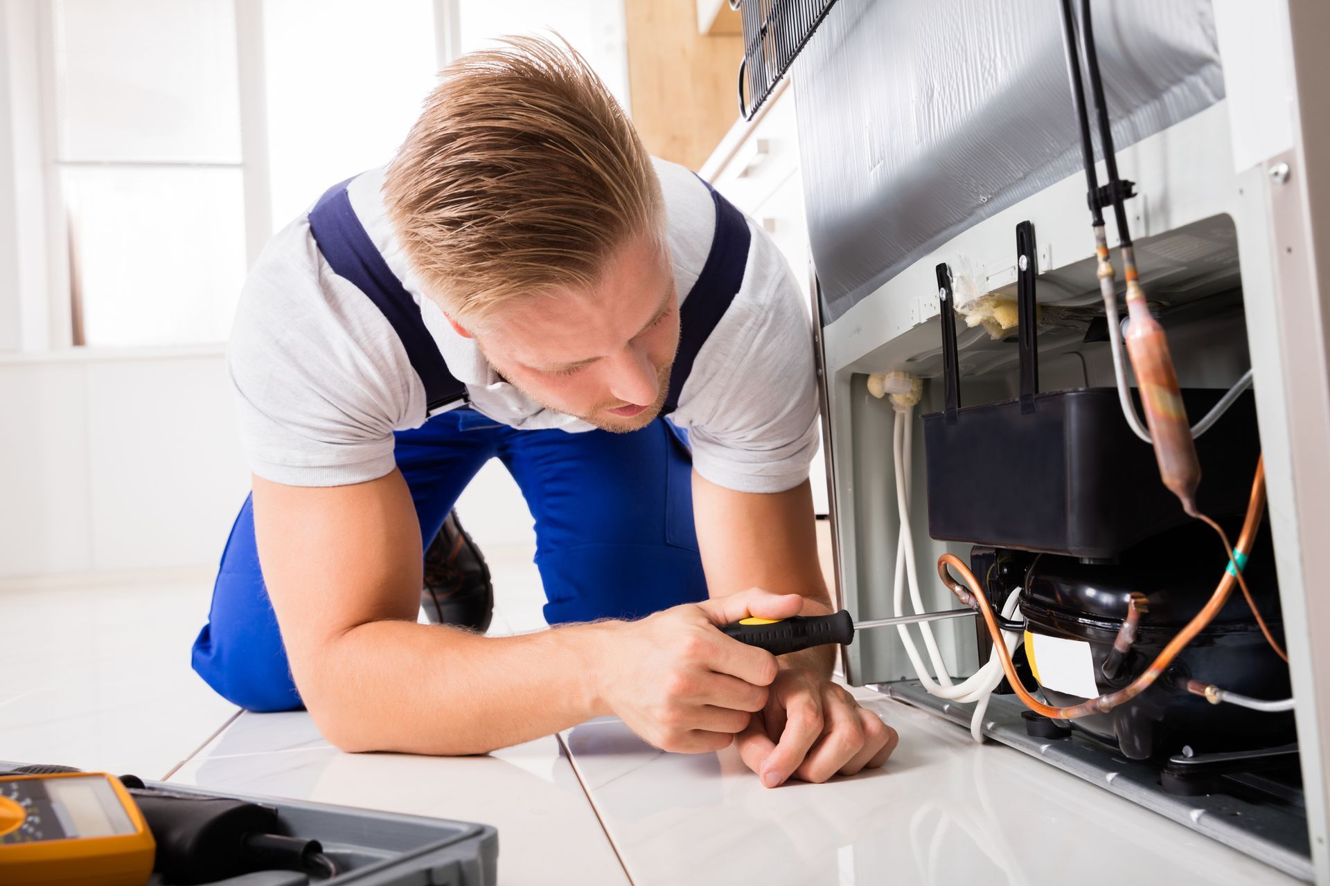 Technician in blue overalls repairs a refrigerator, using a screwdriver.