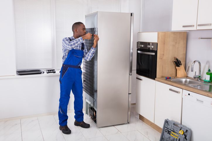 Repairman in blue overalls repairs a refrigerator in a kitchen with white cabinets.
