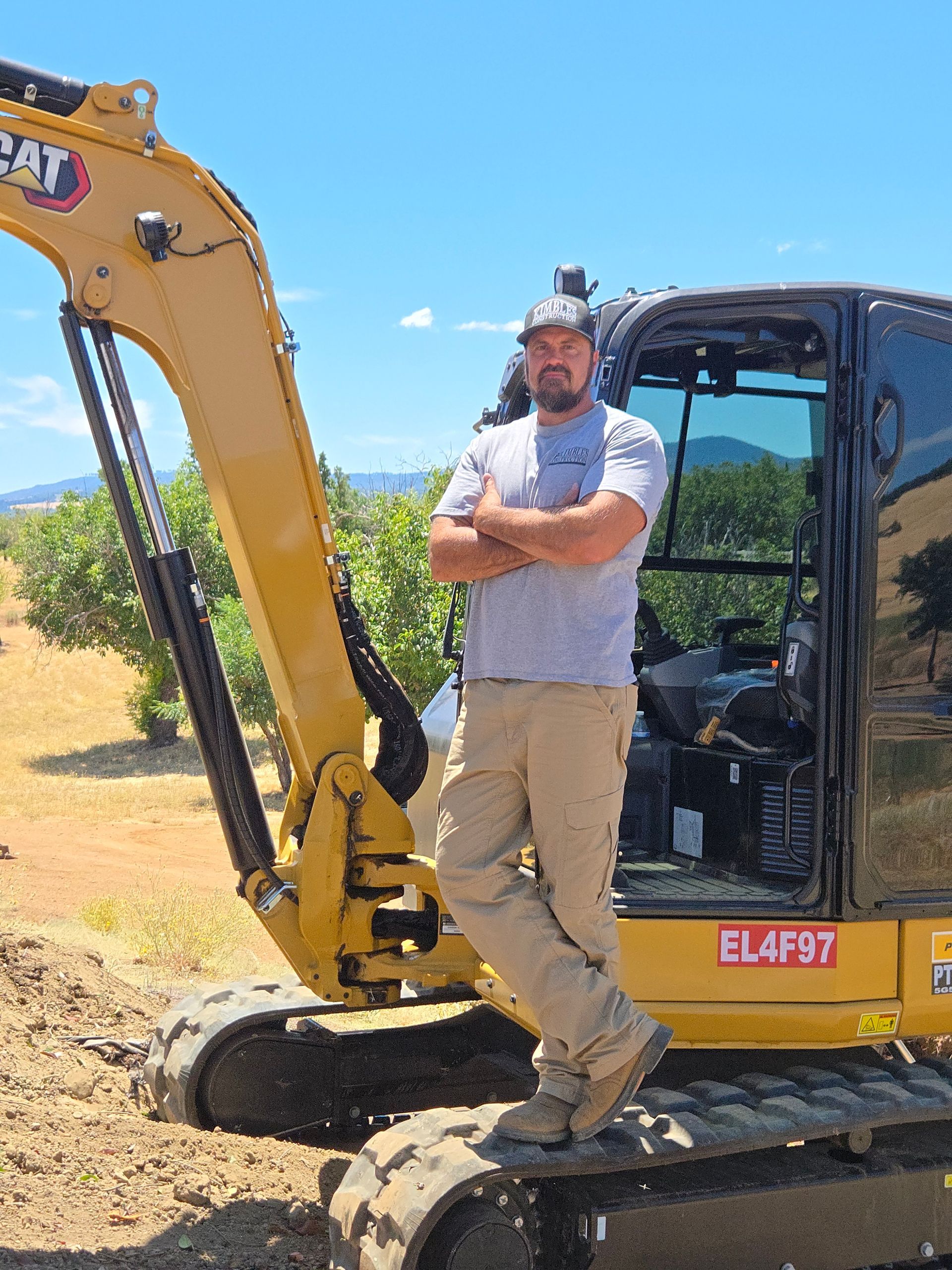 Man in work clothes standing on an excavator, arms crossed, outdoors. Blue sky, yellow machine.