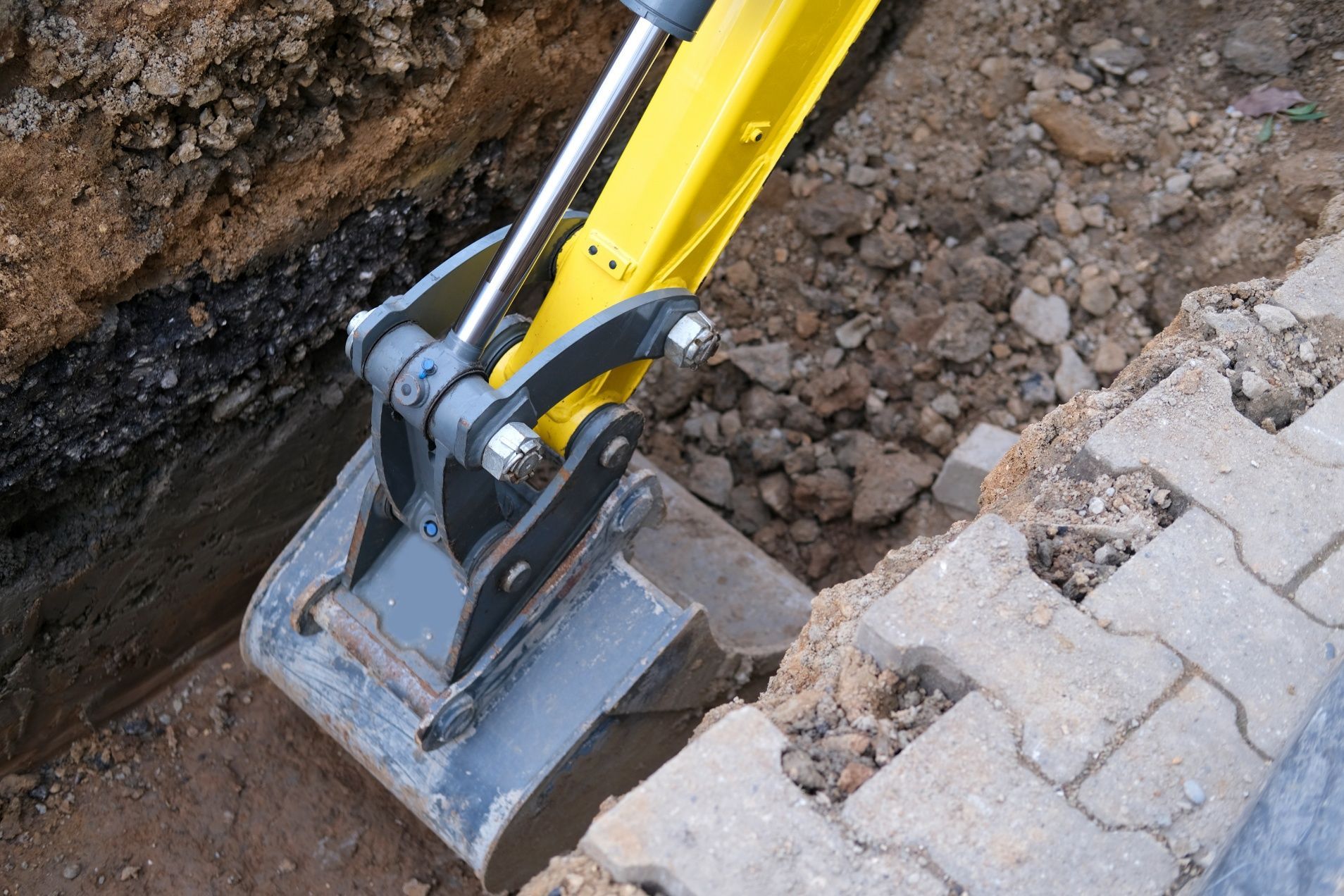 Yellow excavator bucket digging a trench next to brick pavers.