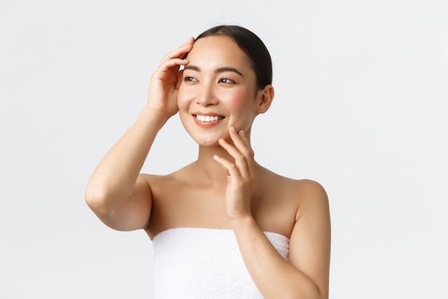 A woman wrapped in a white towel is touching her face and smiling.