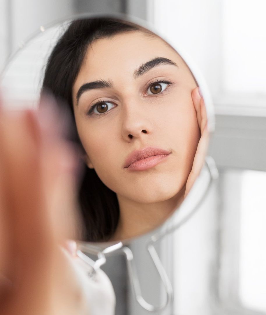 A woman is looking at her face in a mirror.