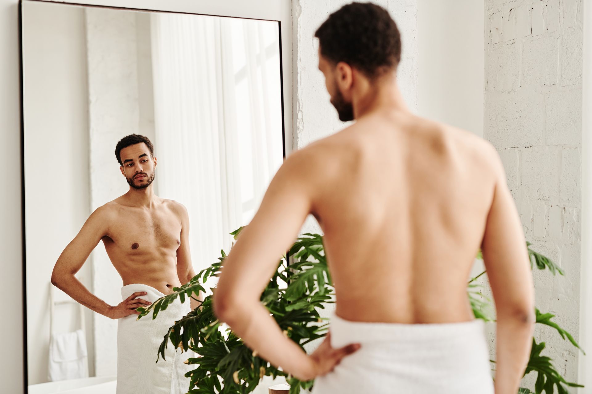 A shirtless man wrapped in a towel is looking at himself in a bathroom mirror.