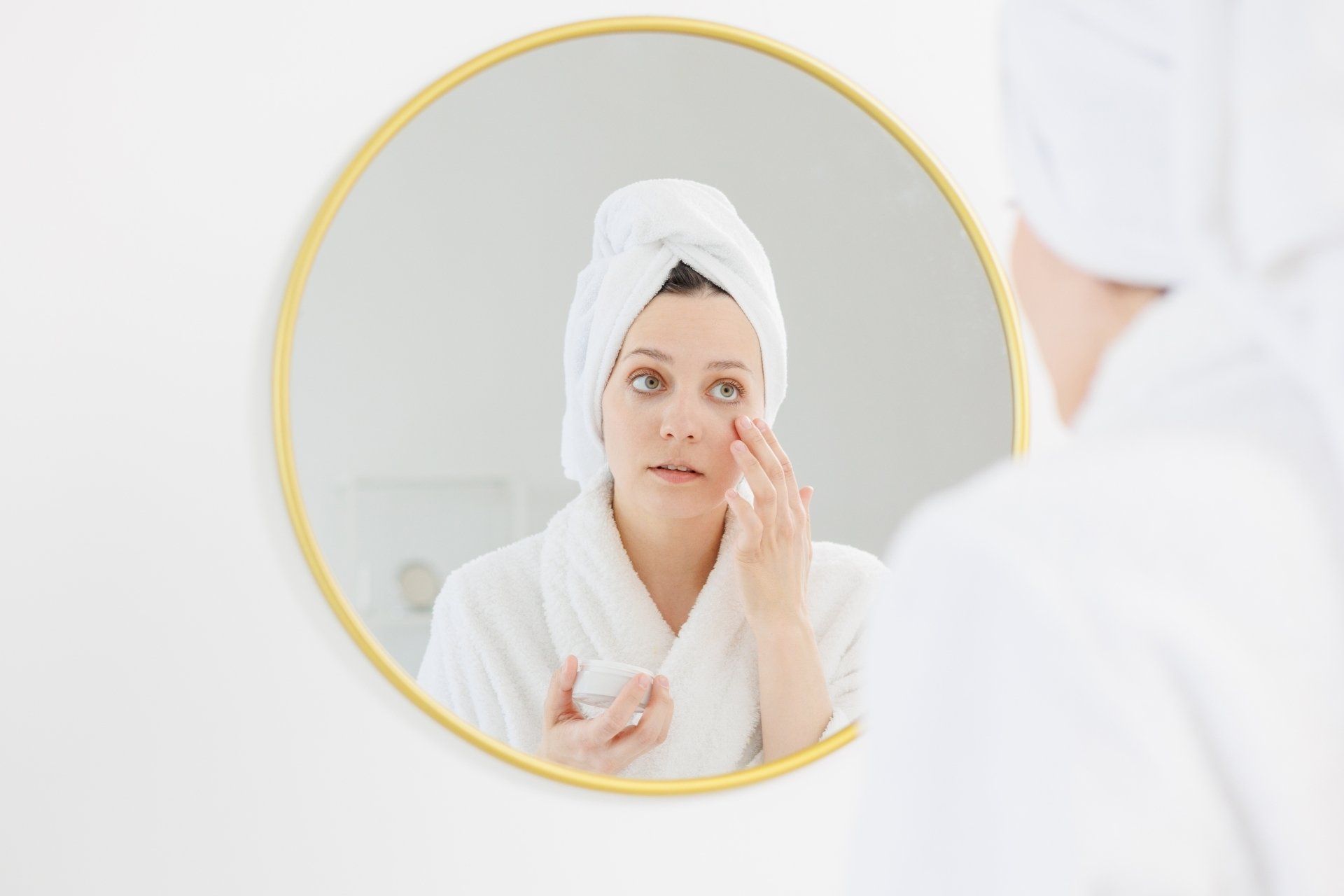 A woman with a towel wrapped around her head is looking at her face in a mirror.