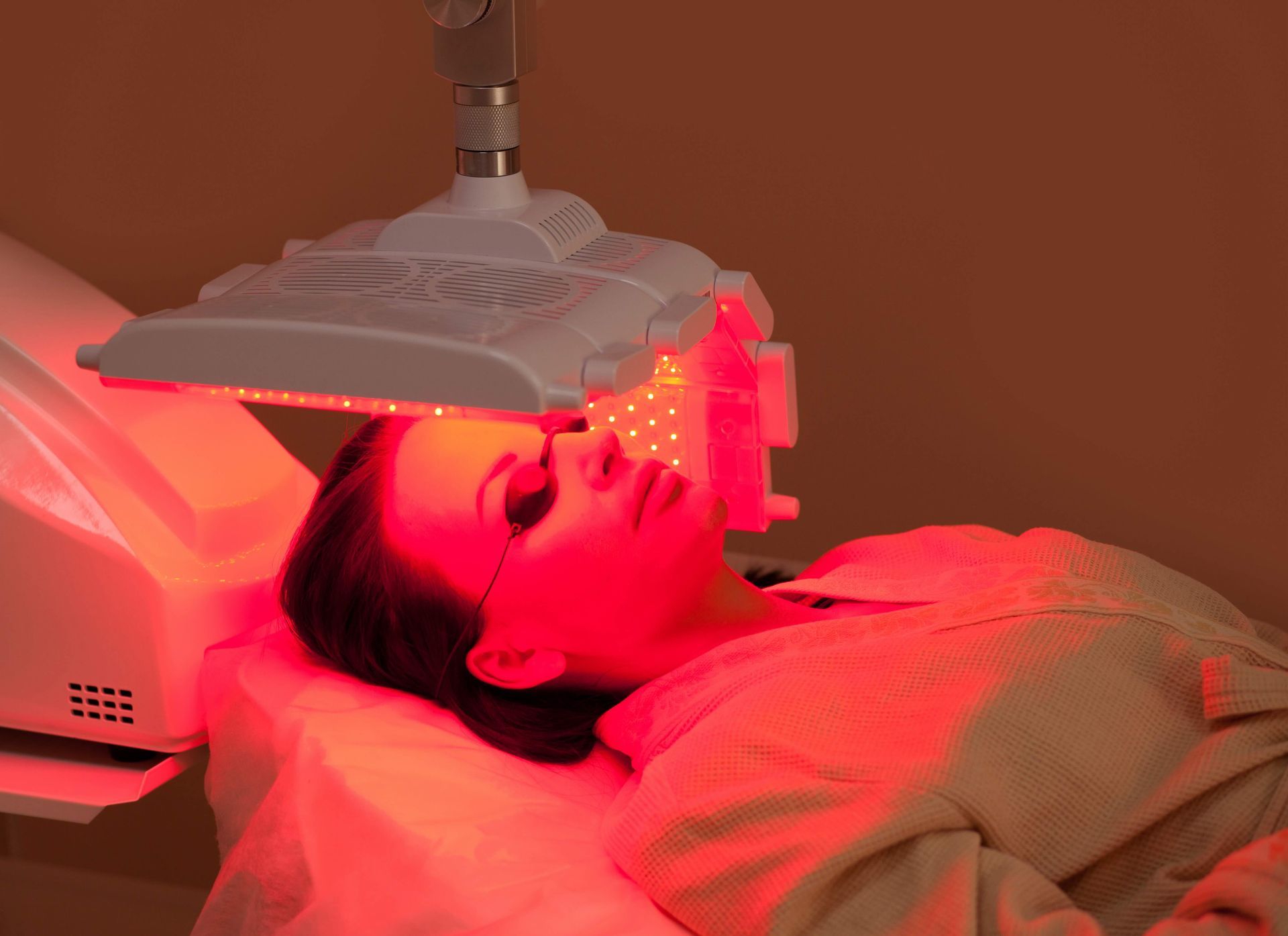 A woman is laying on a bed getting a red light treatment on her face.