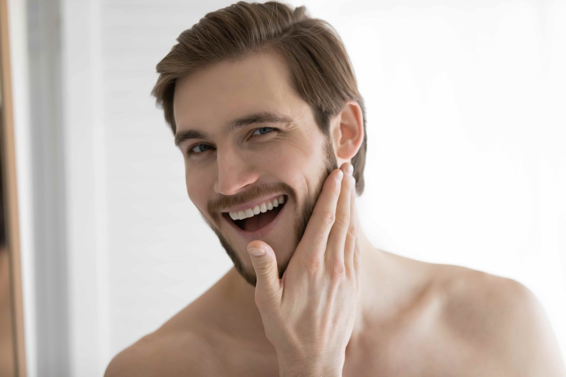 A shirtless man is smiling while looking at his face in the mirror.