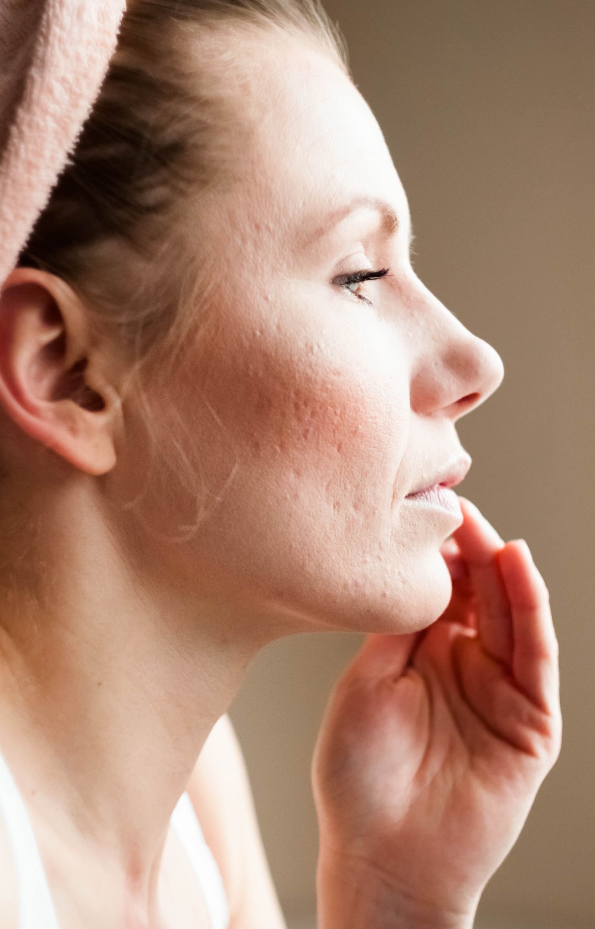 A woman with a towel wrapped around her head is looking at her face in the mirror.