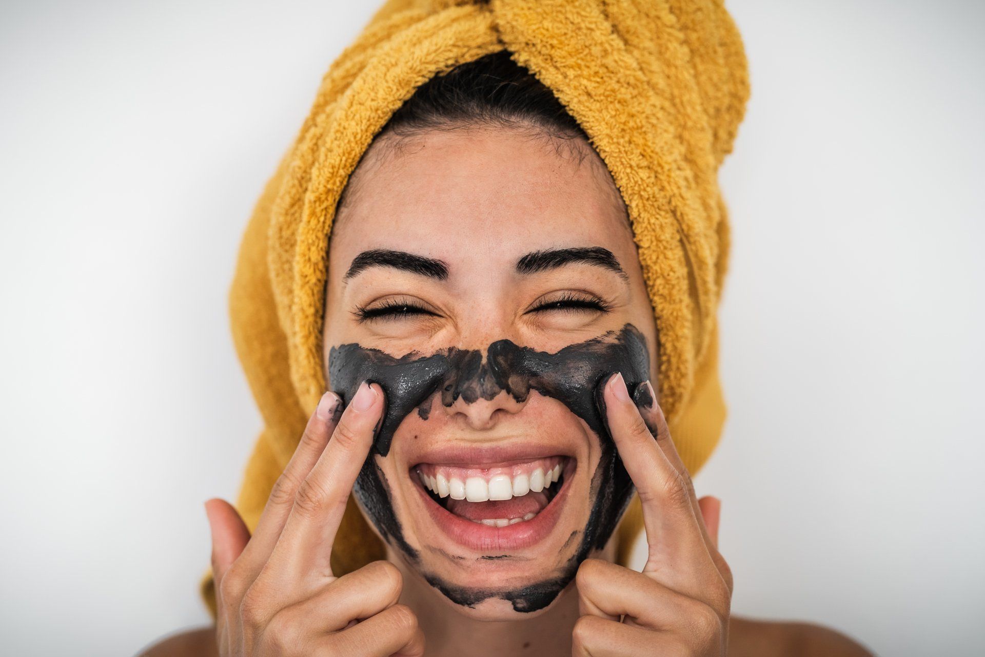 A woman with a towel wrapped around her head is applying a black mask to her face.