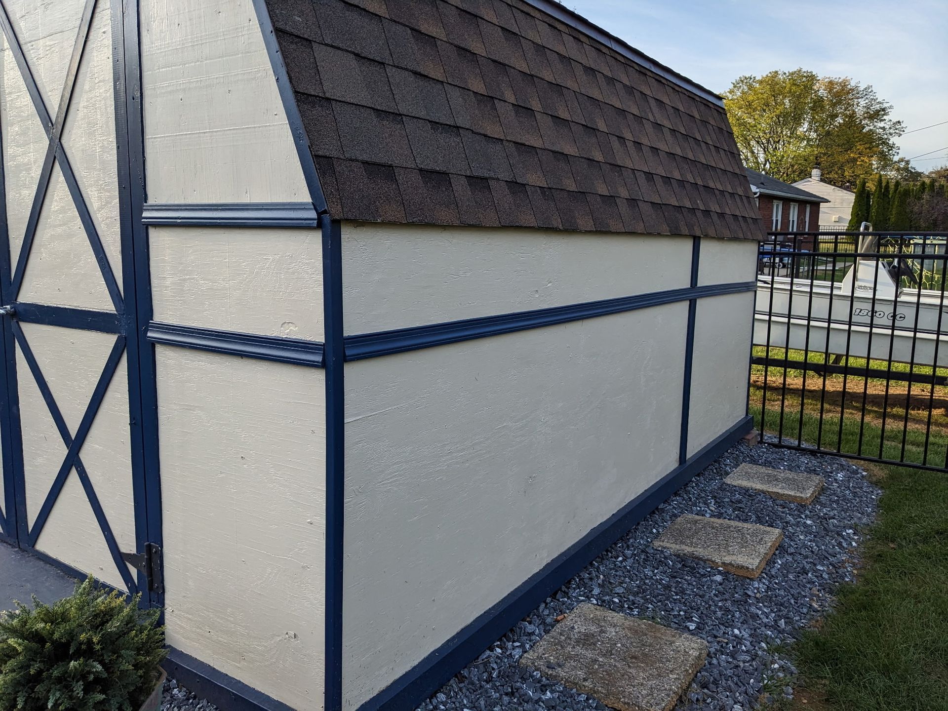 Side view of a shed with a brown shingle roof, cream walls, and blue trim, next to a black fence.
