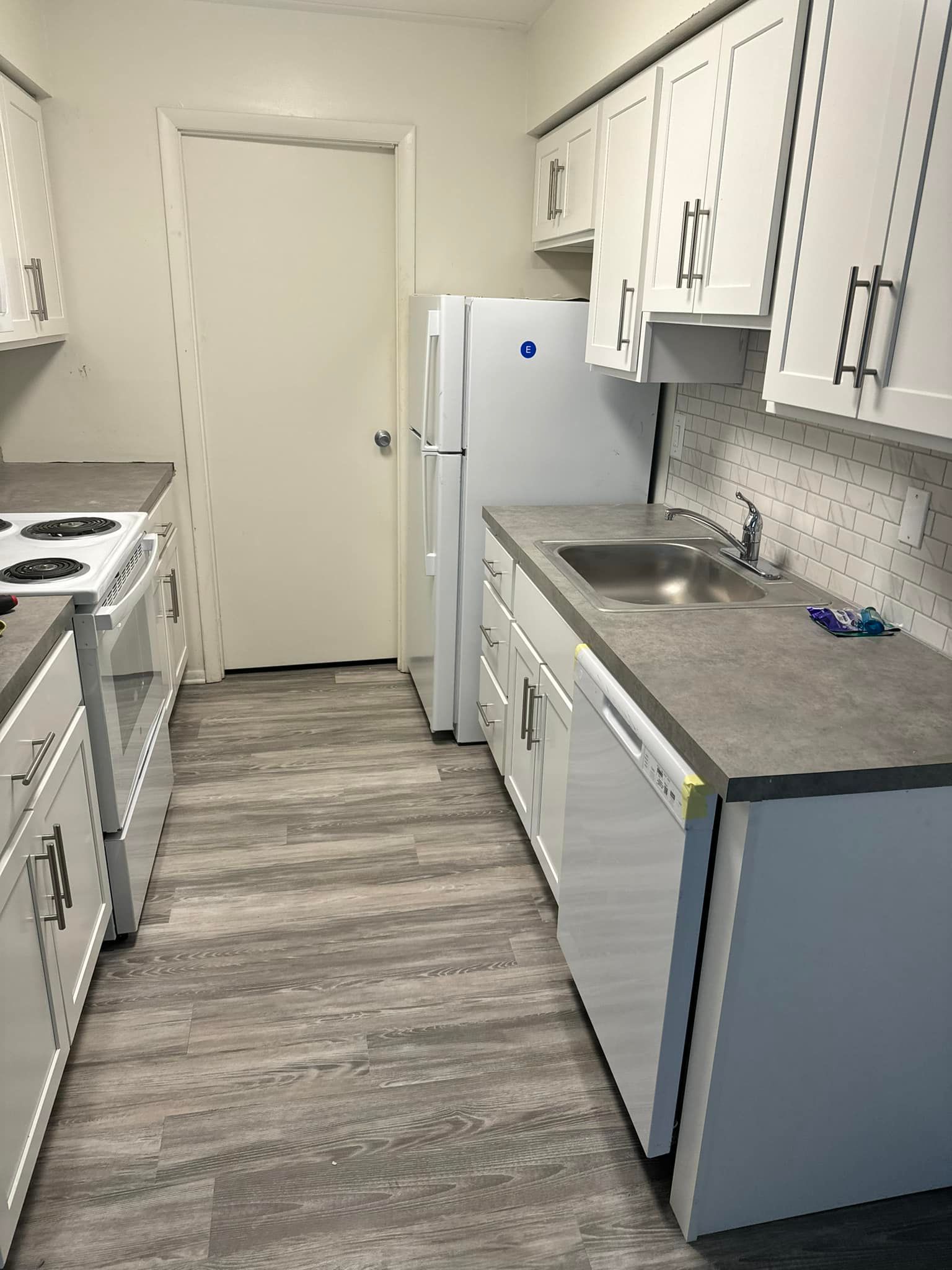 Narrow kitchen with white cabinets, gray countertops, and wood-look flooring. A stove, refrigerator, and sink are visible.