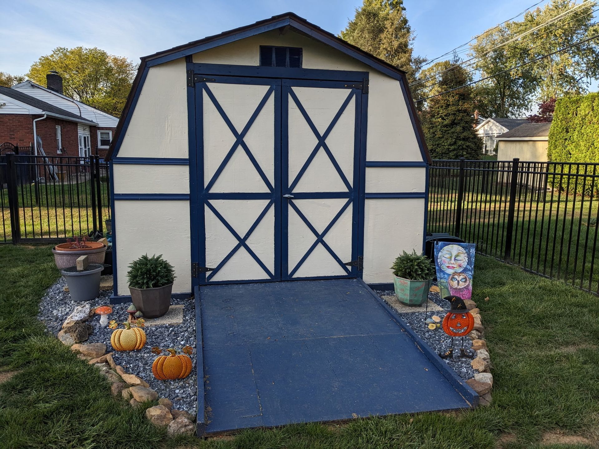 Shed with blue ramp, doors, and trim. Decorated with pumpkins and plants. Black fence, trees, and grass.