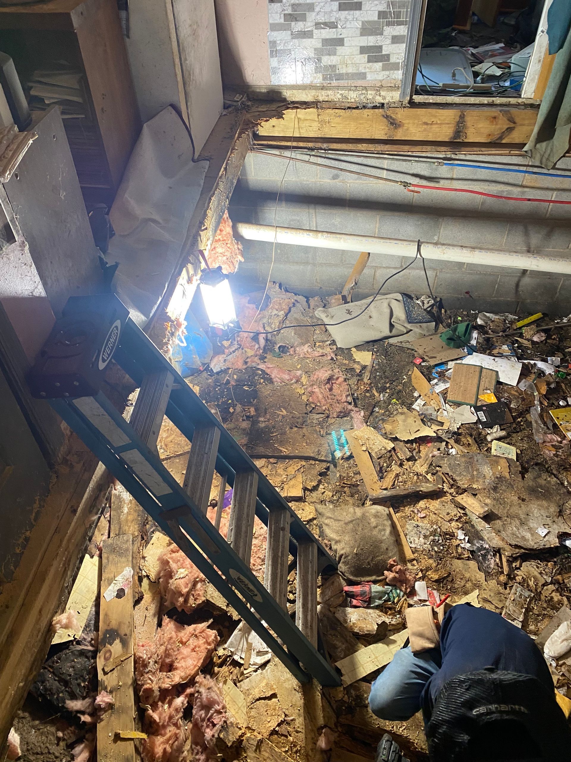 Ladder in a damaged ceiling. A person works in a cluttered space with exposed insulation, wood, and debris.