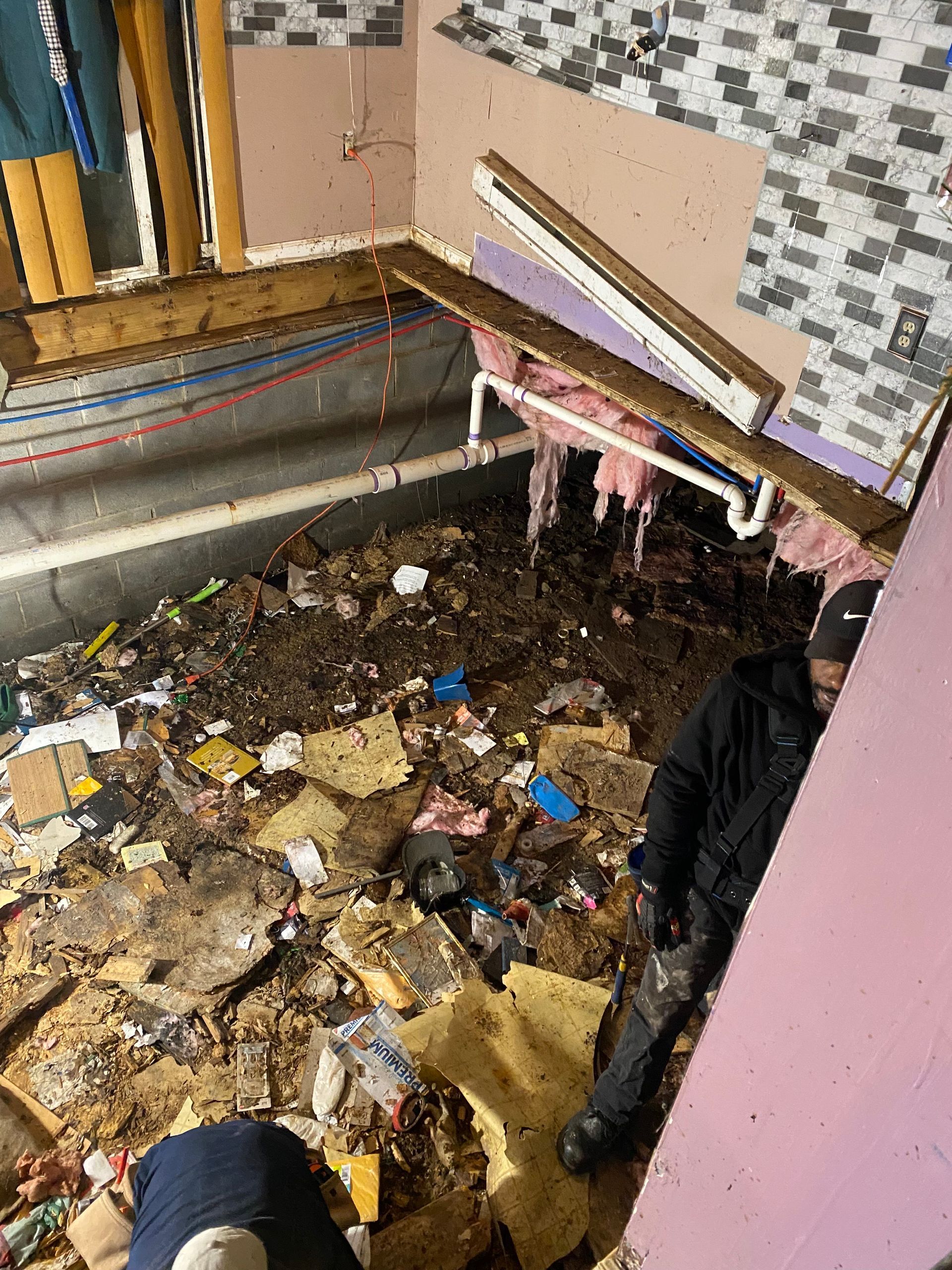Renovated bathroom debris and person in dark clothes. Pink wall, white pipes, exposed insulation, dirty floor.