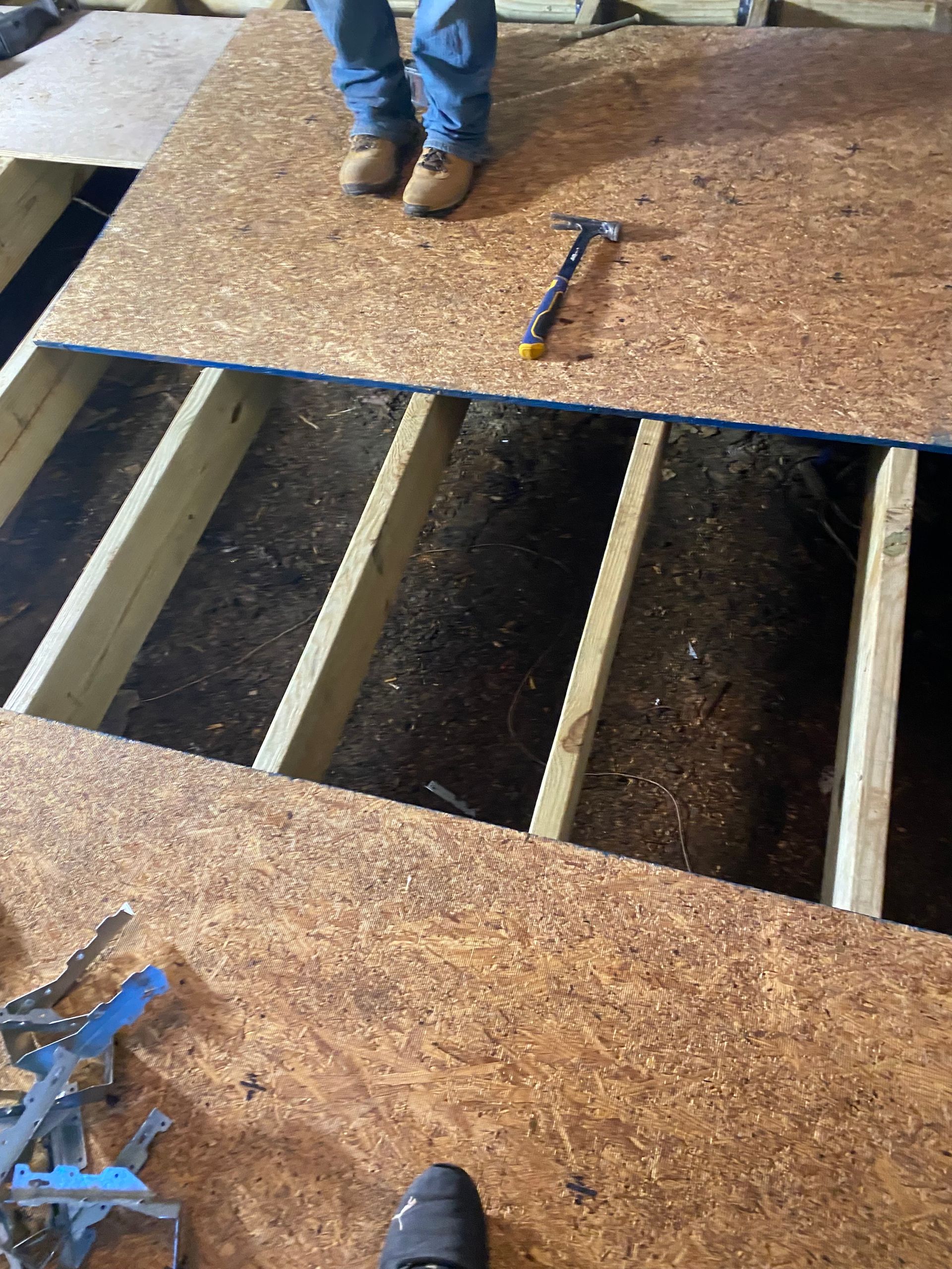 Person standing near an opening in a particle board floor exposing floor joists.