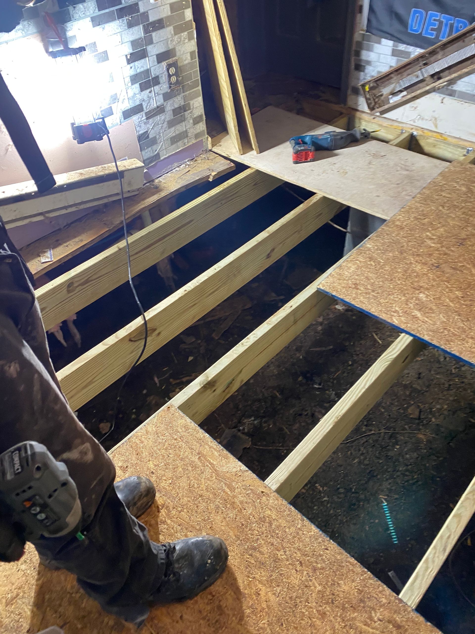 Person installing a wood floor; plywood sheets laid over exposed joists in a room with a light fixture and tool in view.