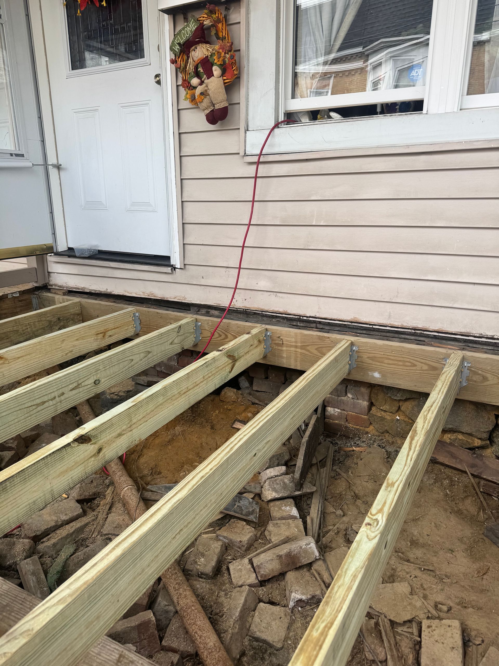 Construction of a deck; wooden beams are installed over old bricks, near a house with a white door.