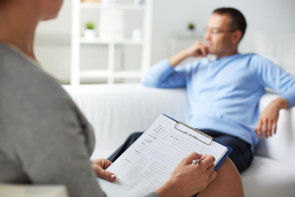 A man is sitting on a couch talking to a woman who is holding a clipboard — Tuncurry Medical Centre In Tuncurry, NSW