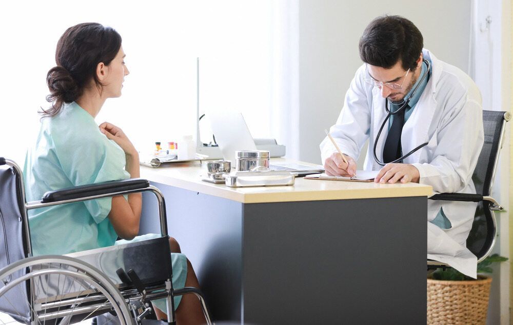 A Woman in A Wheelchair Is Sitting at A Desk Talking to A Doctor — Tuncurry Medical Centre In Tuncurry, NSW
