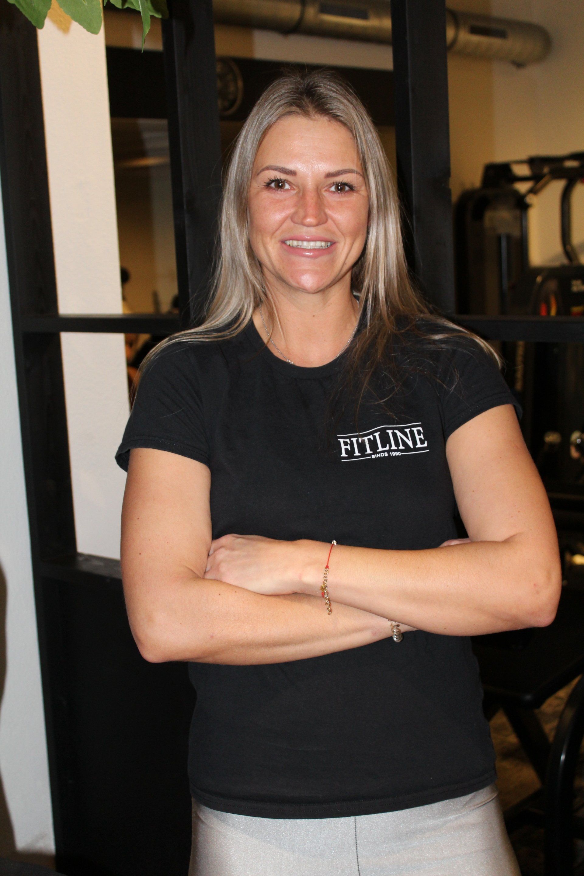 Een vrouw in een zwart t-shirt staat met haar armen over elkaar in een sportschool.