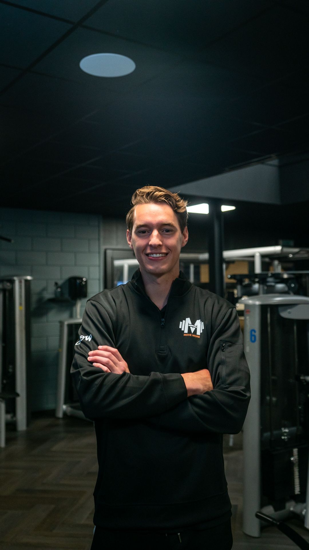 Een vrouw in een zwart shirt staat in een sportschool met haar handen in haar zij.