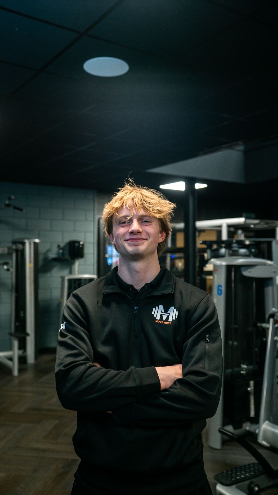 Een vrouw in een zwart shirt staat met haar armen over elkaar in een sportschool.
