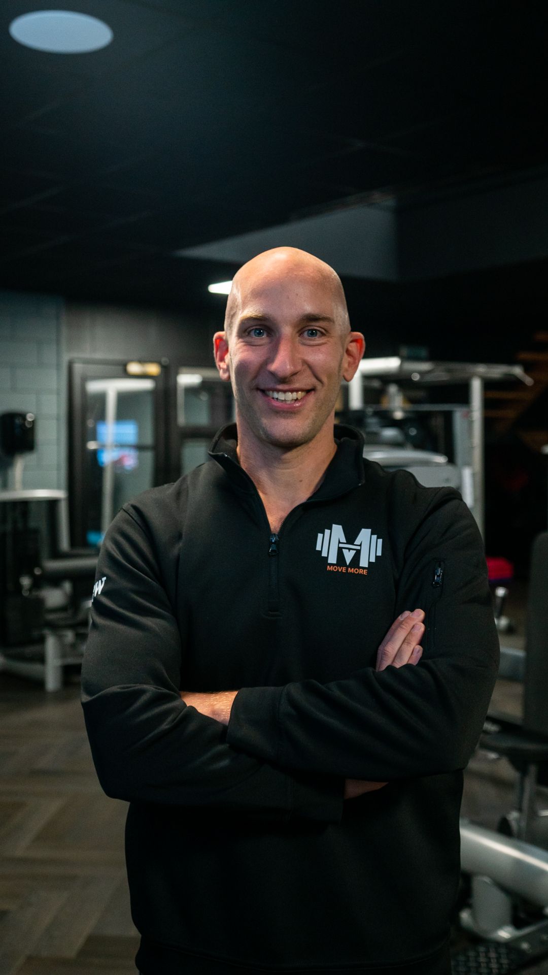 Een man in een zwart shirt staat met zijn armen over elkaar in een sportschool.