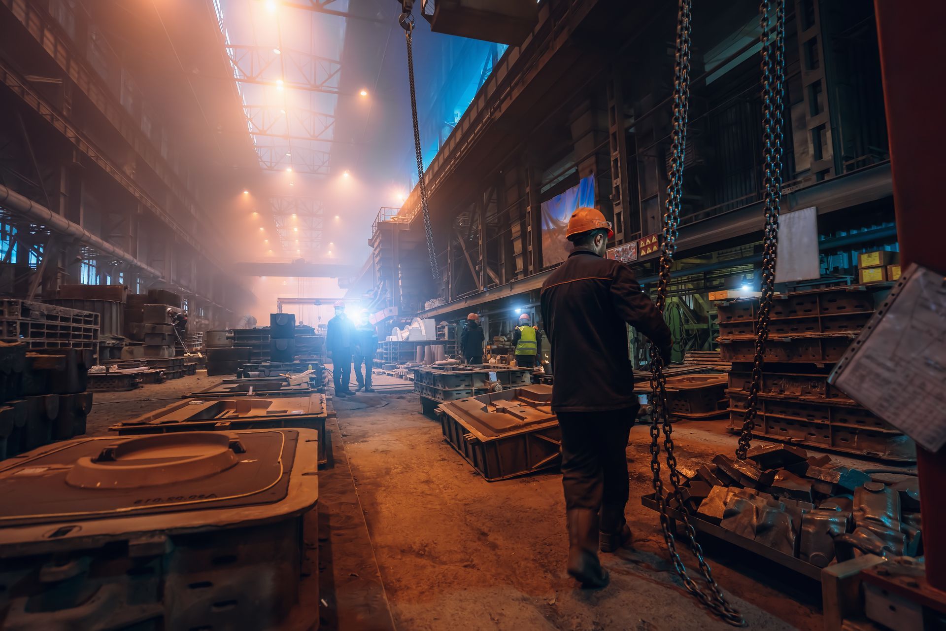 Worker in hard hat walks through a dimly lit industrial factory, surrounded by metal molds and machinery.