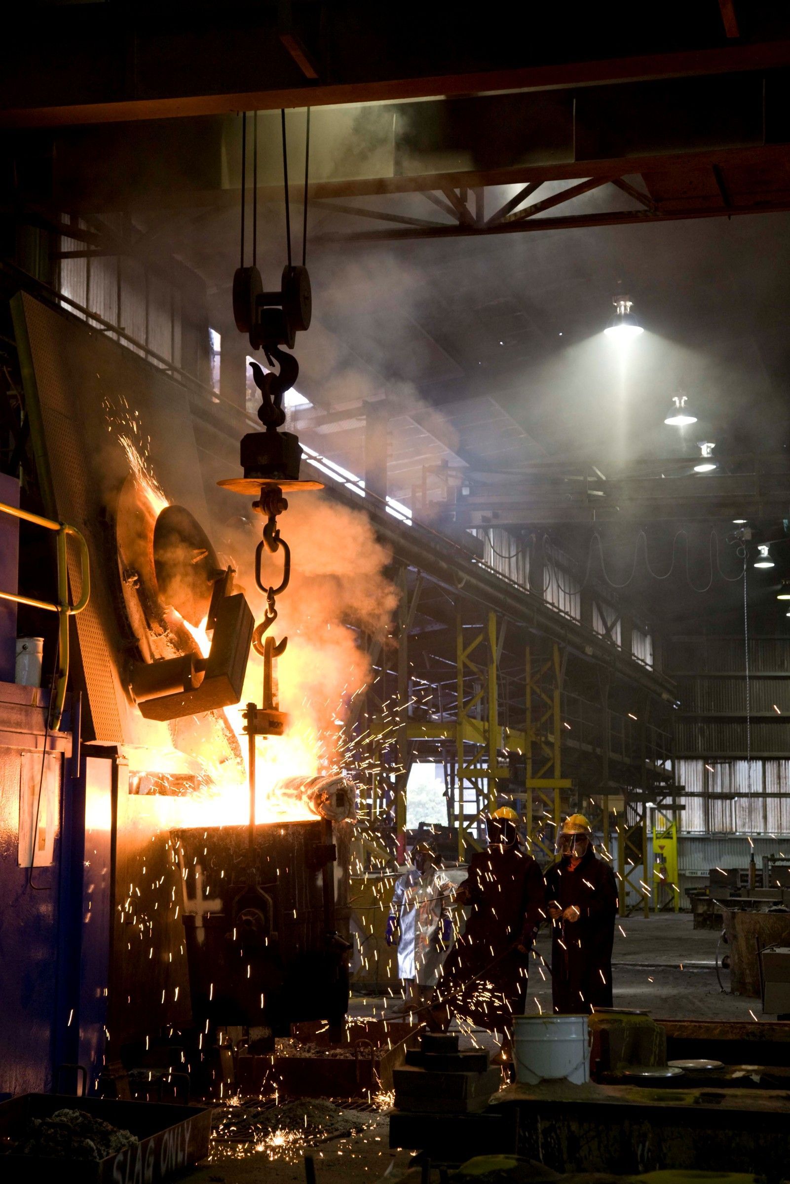Molten metal being poured in a factory, creating sparks and smoke. Workers in safety gear observe.
