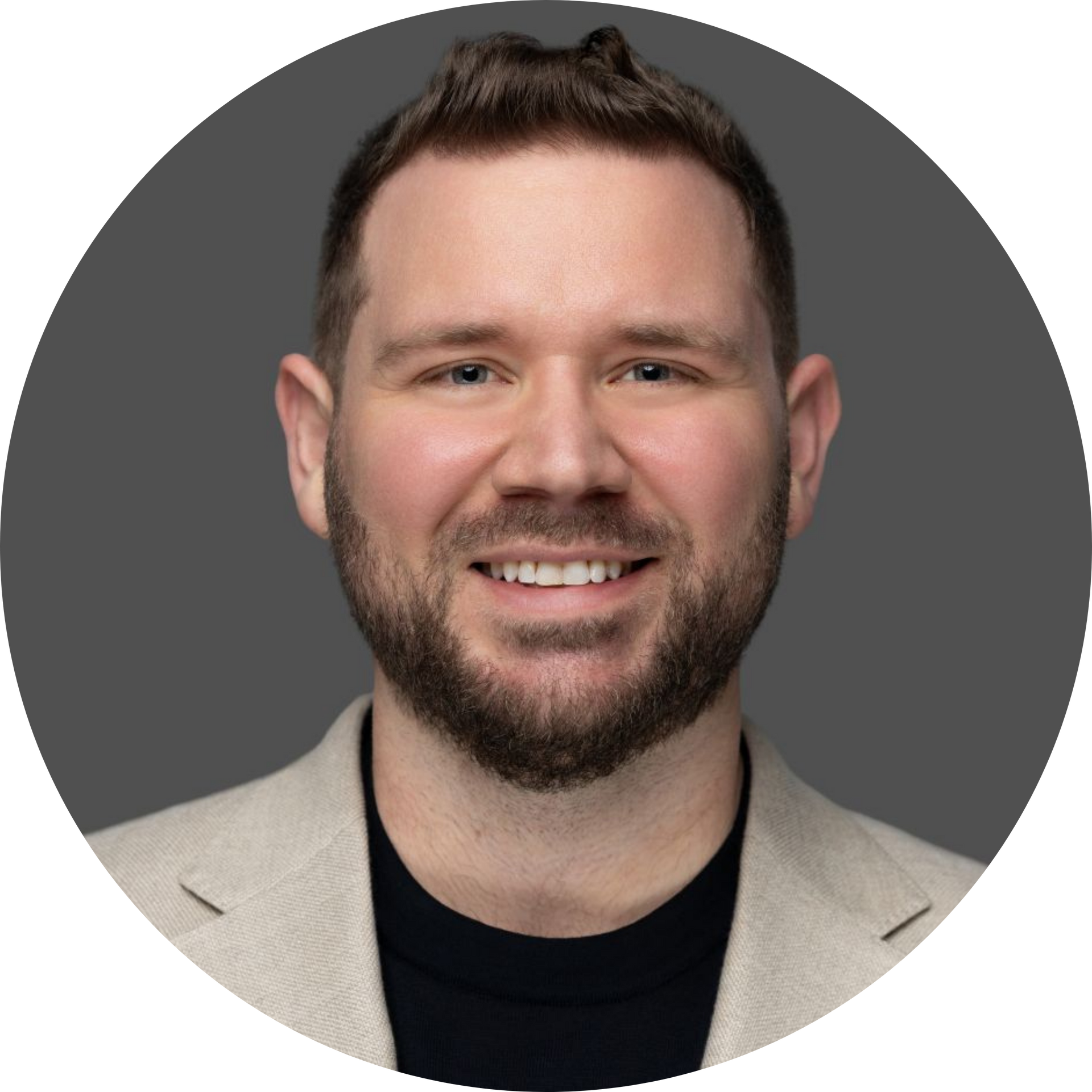 Man with a short beard smiles, wearing a black shirt and tan blazer, against a gray background.