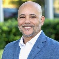 A smiling person with shoulder-length blonde hair, wearing a black shirt and a silver cross necklace against a white backdrop.