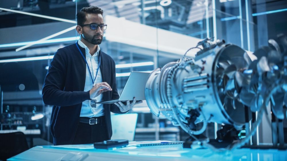 Man with glasses using a laptop in a laboratory, examining a large metallic engine.