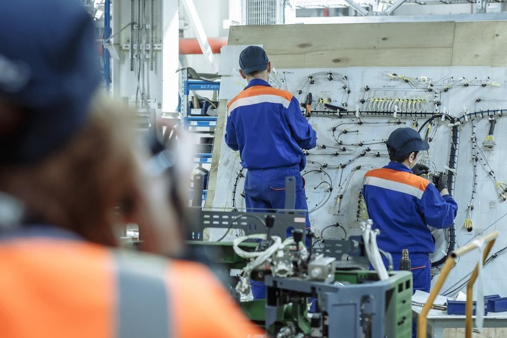 Workers in blue uniforms assemble wiring on a large panel in a factory.