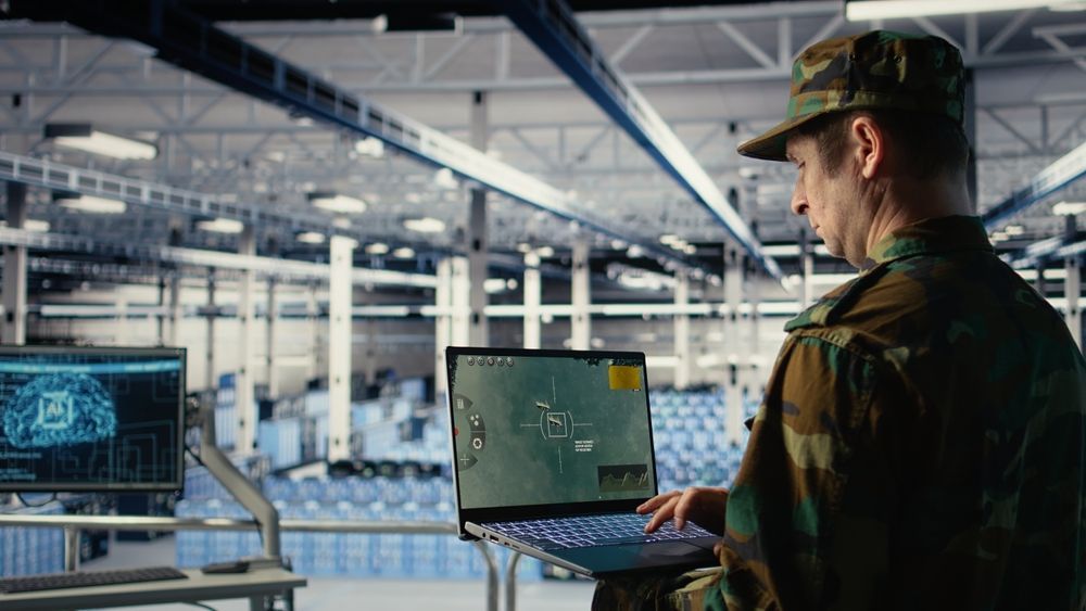 A person in camouflage operates a laptop with a map overlay, in a large, industrial facility.