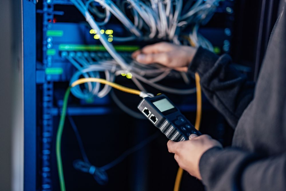 Person using a handheld device to troubleshoot network cables in a server room, with blue lights and wires.