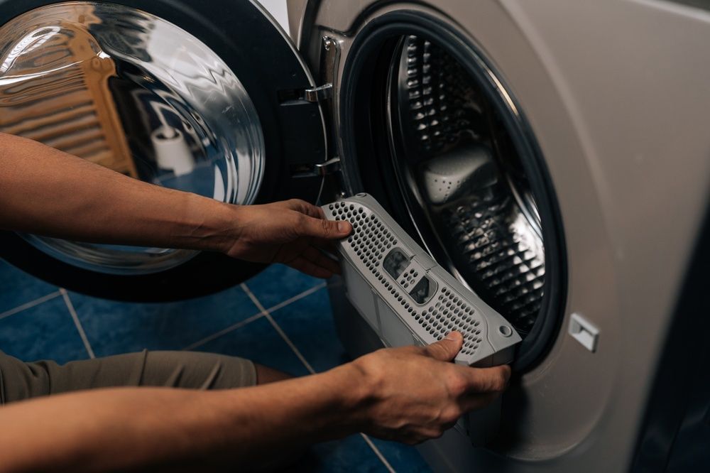 A person removes a gray lint filter from the drum of an open front-load washing machine.