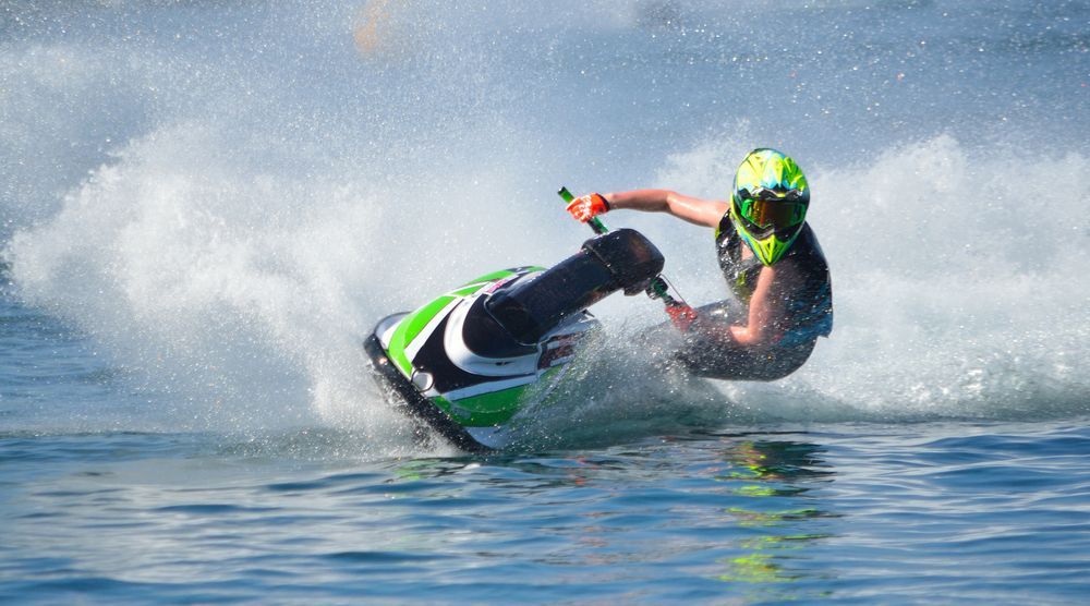 A person on a jet ski, leaned over, spraying water. The rider wears a helmet and life vest on the water.
