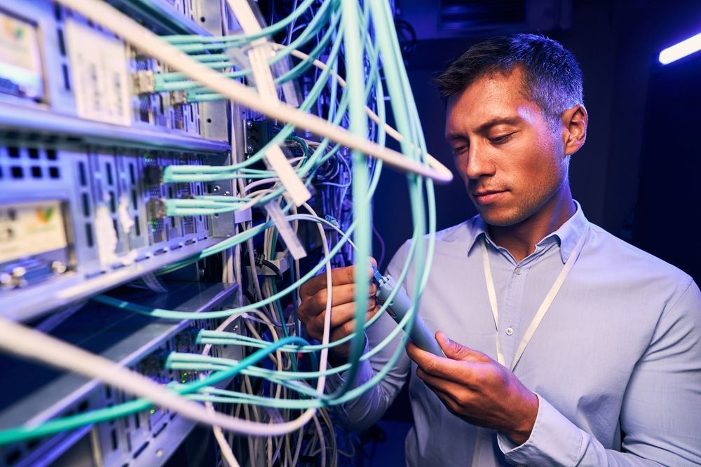 Man examining cables in a server room; he wears a light blue shirt.