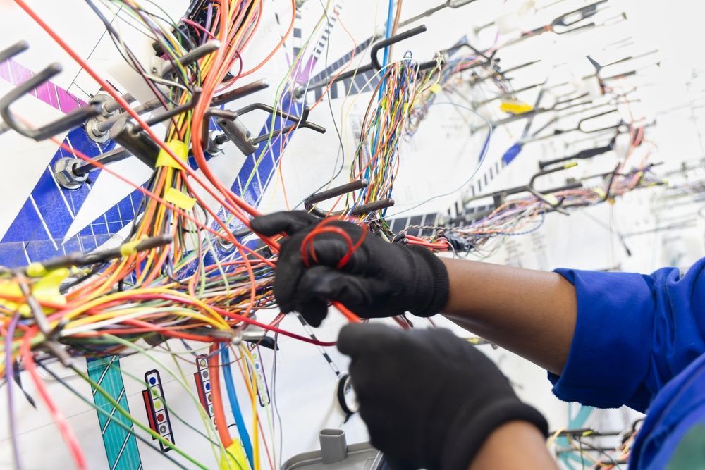 Hands in black gloves working on a complex wiring assembly, with many colored wires.